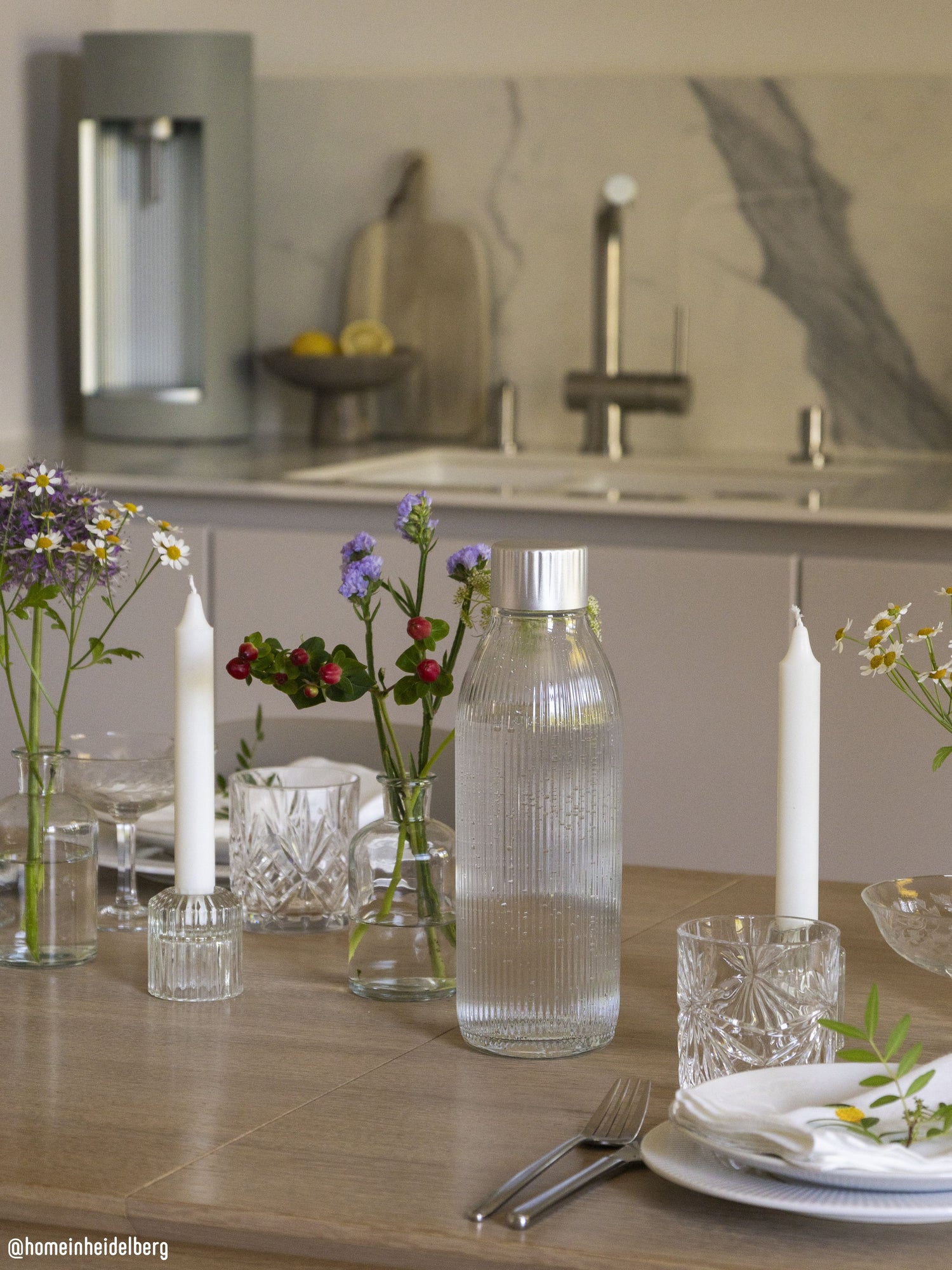 Mysoda glass bottle on a dinner table shet with white tableware, glass candle holders and vases set with small wildflowers. A green Glassy sparkling water maker is visible on the kitchen counter in the background