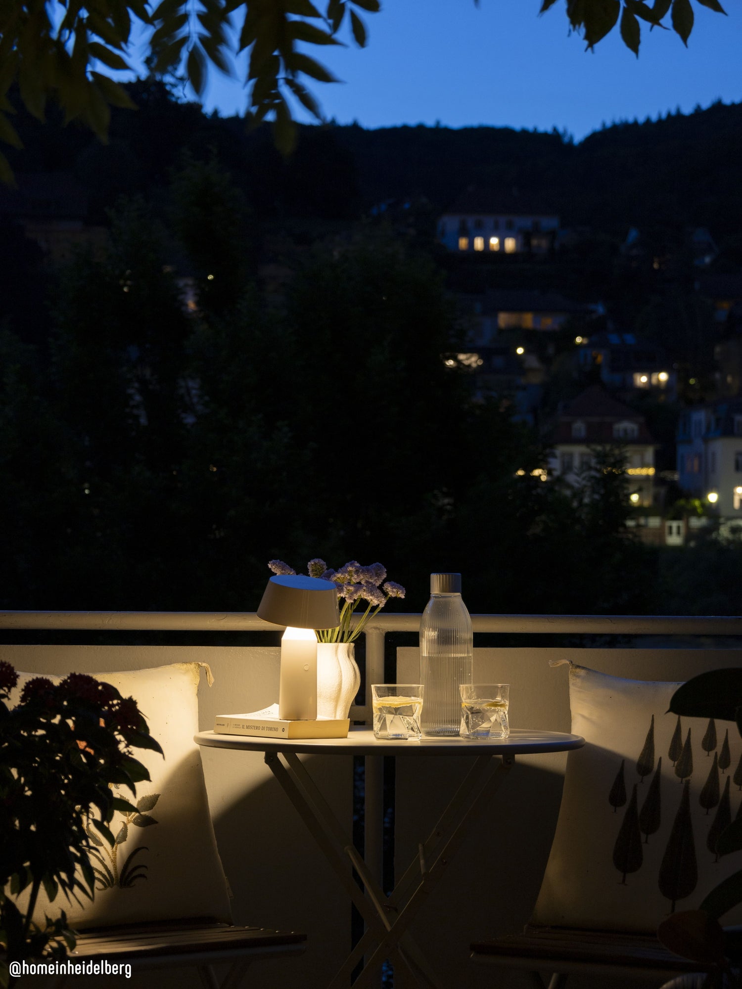 Mysoda glass bottle and glassws of fresh sparkling water illuminated by a lone table lamp on a terrace table. Background gives view over a city by night