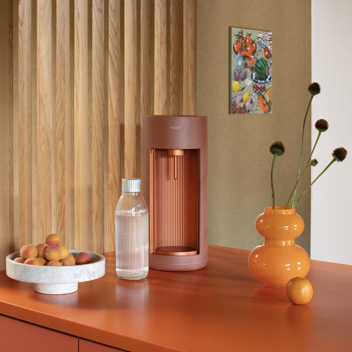 A terracotta-coloured Glassy sparkling water maker and glass bottle on an orange table with matching fruit and flower decoration to the sides
