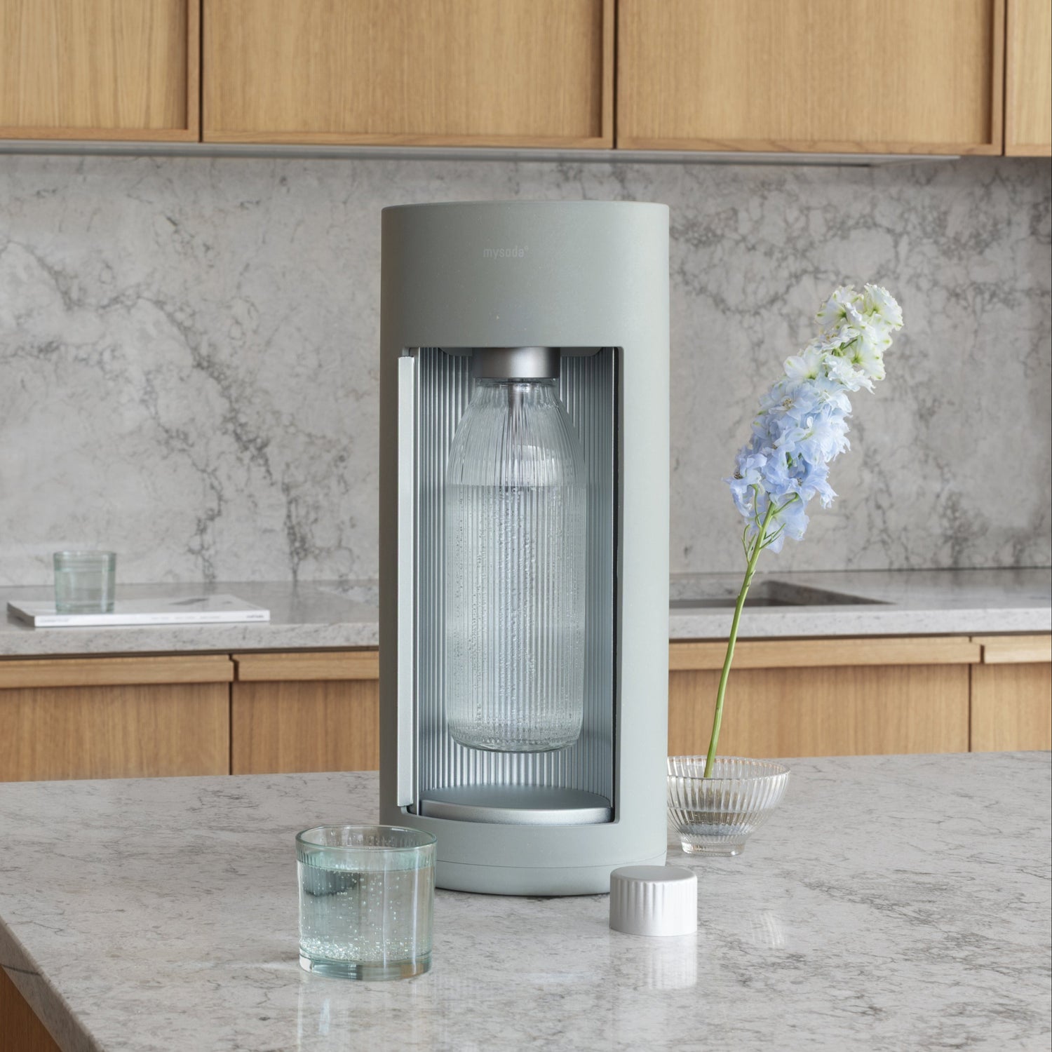 A pigeon coloured Glassy soda maker in a kitchen with gray marble counters and cupboards in light wood