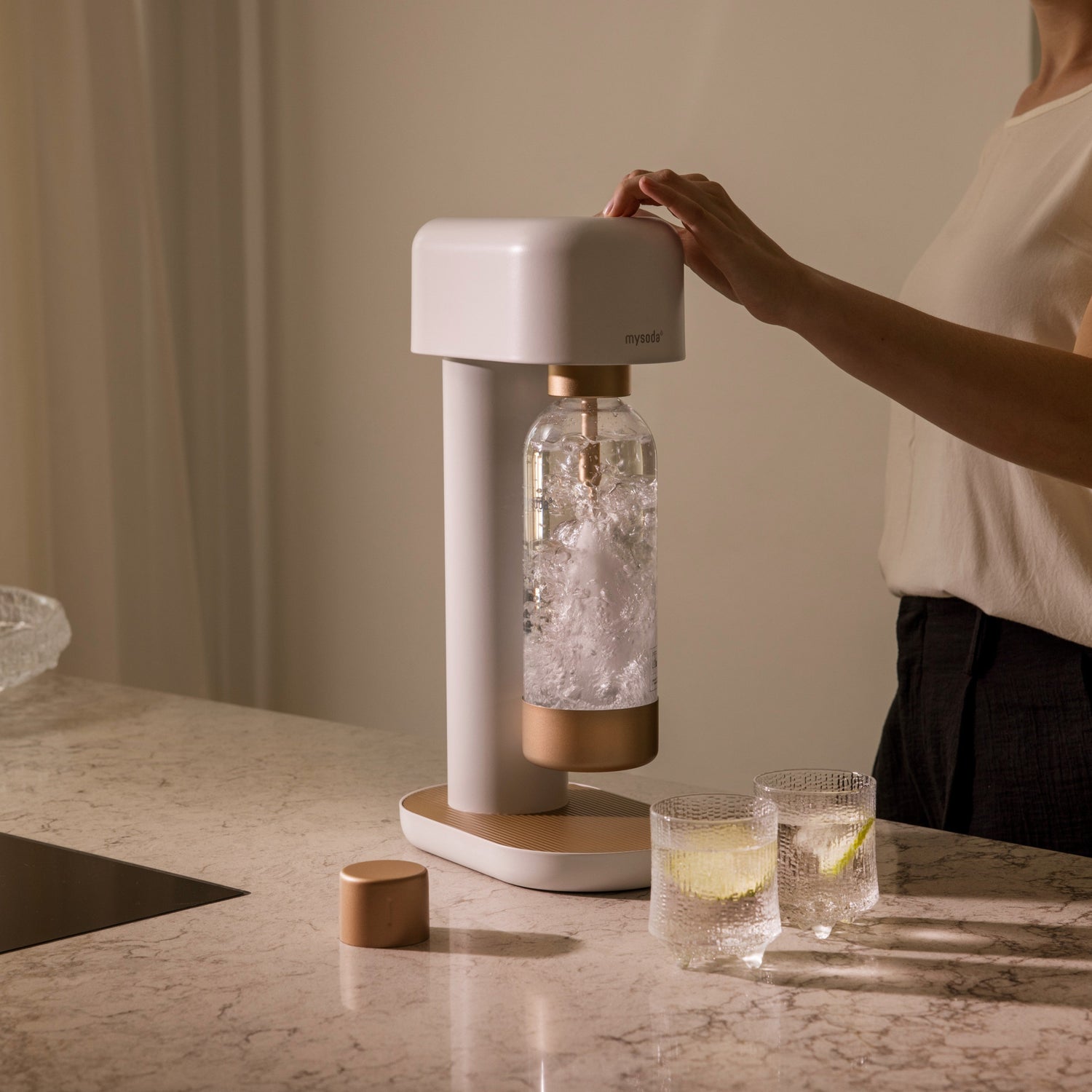 A person carbonating water with a white-copper Ruby 2 sparkling water maker that is standing on a marble counter