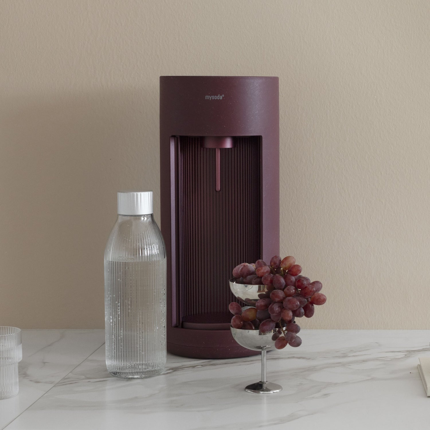A beetroot Glasssy sparkling water maker with glass bottles and a bowl of fresh grapes