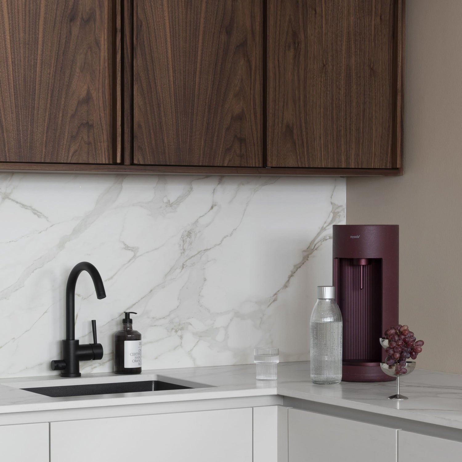A beetroot-coloured Glassy soda maker with glass bottle in a kitchen setting in white marble and dark wood