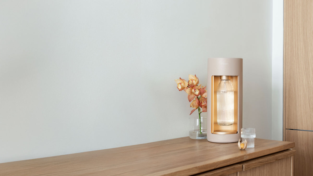 A Glassy sparkling water maker, colourlatte, and matching flower decoration on a wooden counter in front of a plain white wall