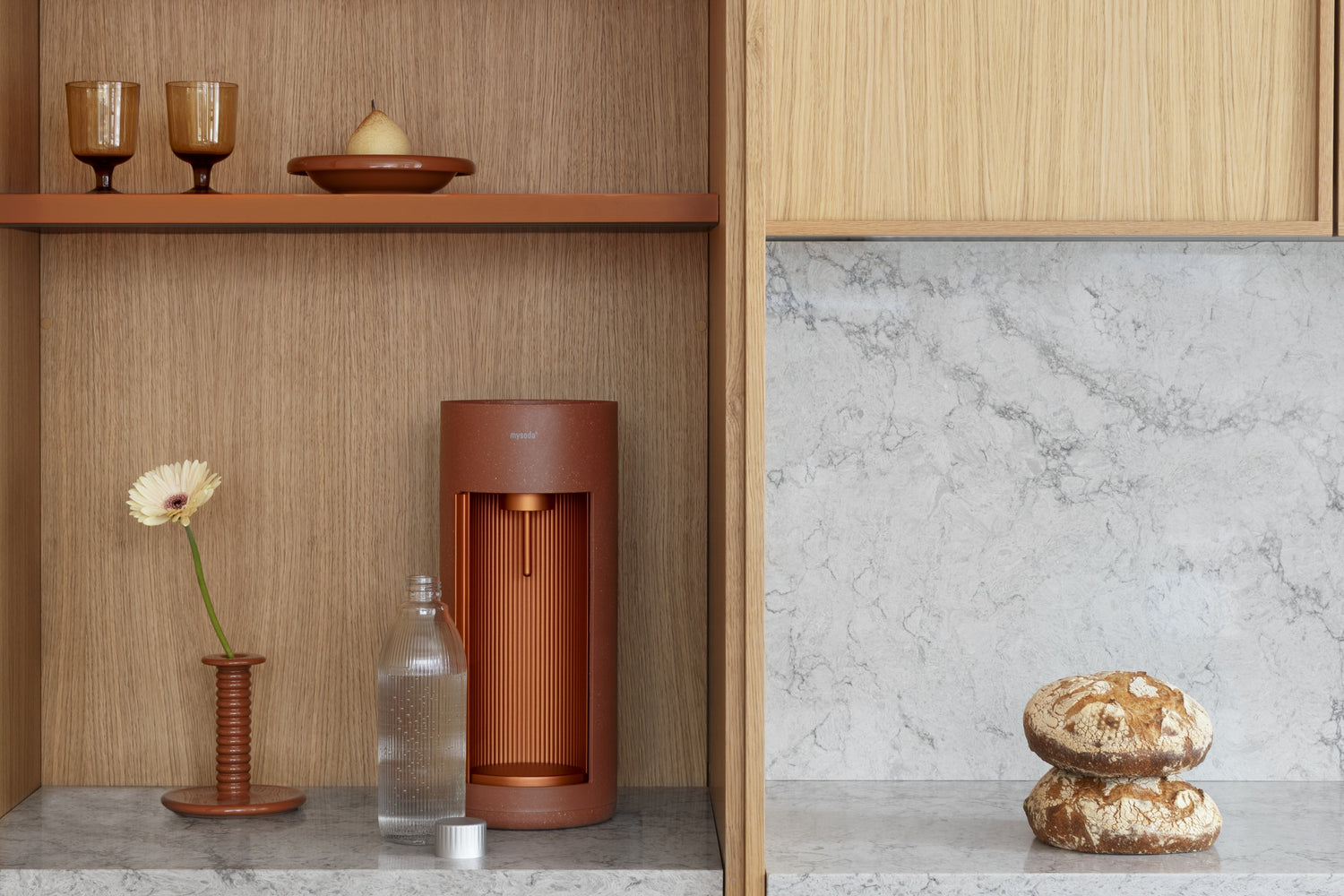 A Glassy sparkling water maker, colour Terracotta, in an open kitchen cabinet af light wood.