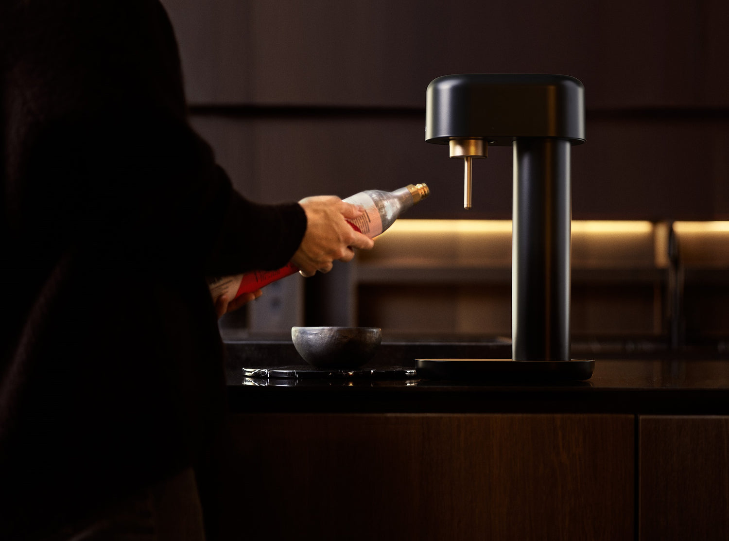 Someone holding a Mysoda CO2 cylinder in front of a black-copper Ruby 2 sparkling water maker in a dark kitchen