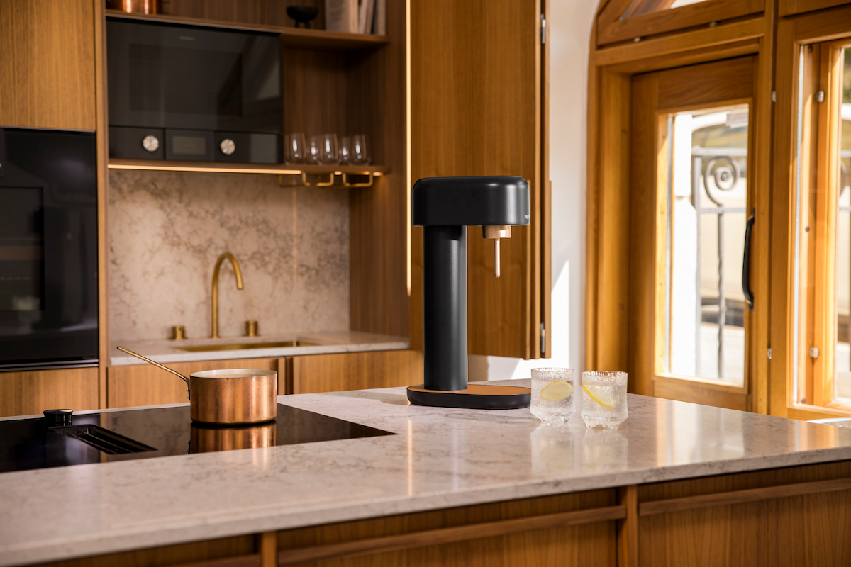 A black-copper Ruby 2 sparkling water maker and two glasses on the countertop in front of opened wooden kitchen cabinets
