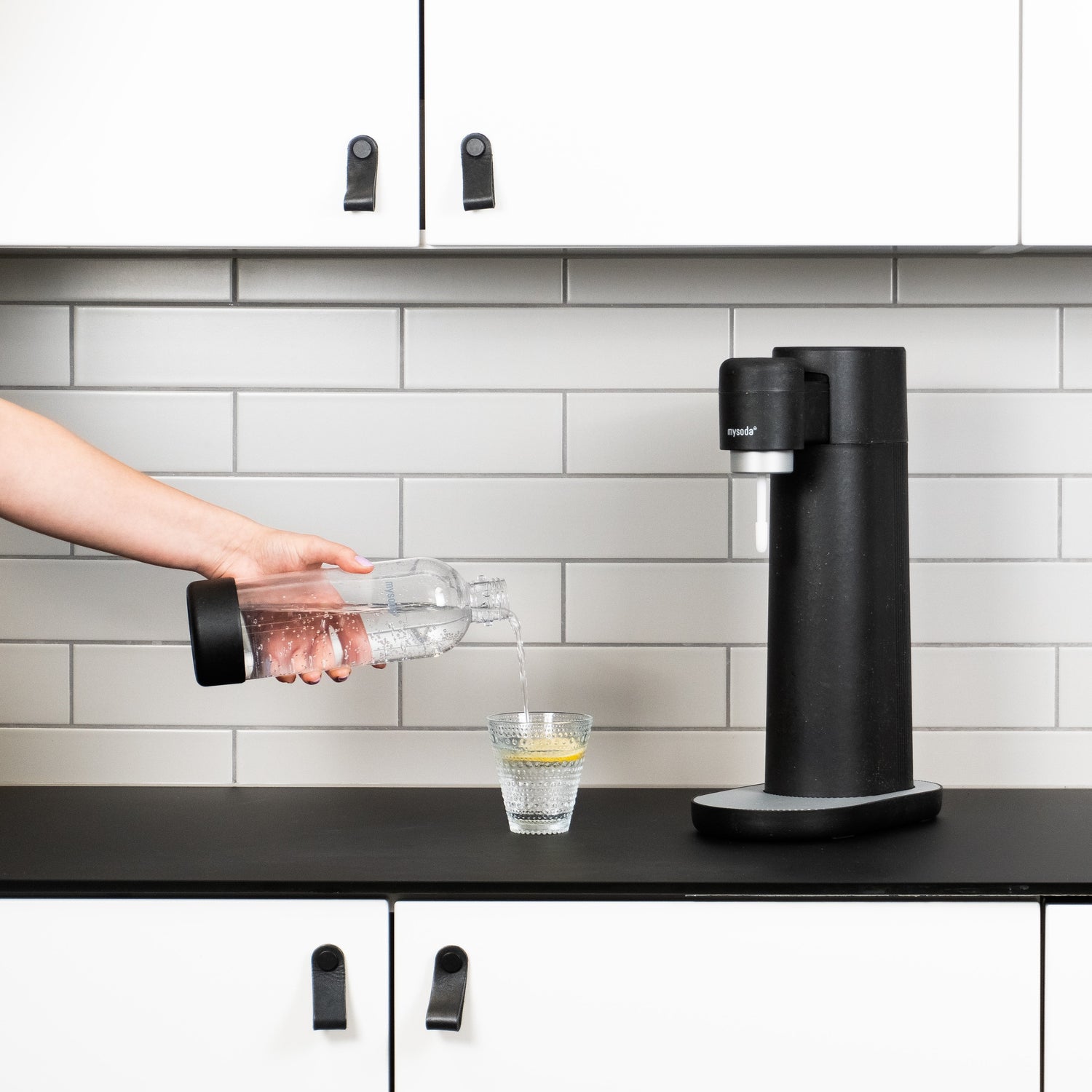 A black Mysoda Toby sparkling water maker in a kitchen next to which someone is pouring a glas of sparkling water