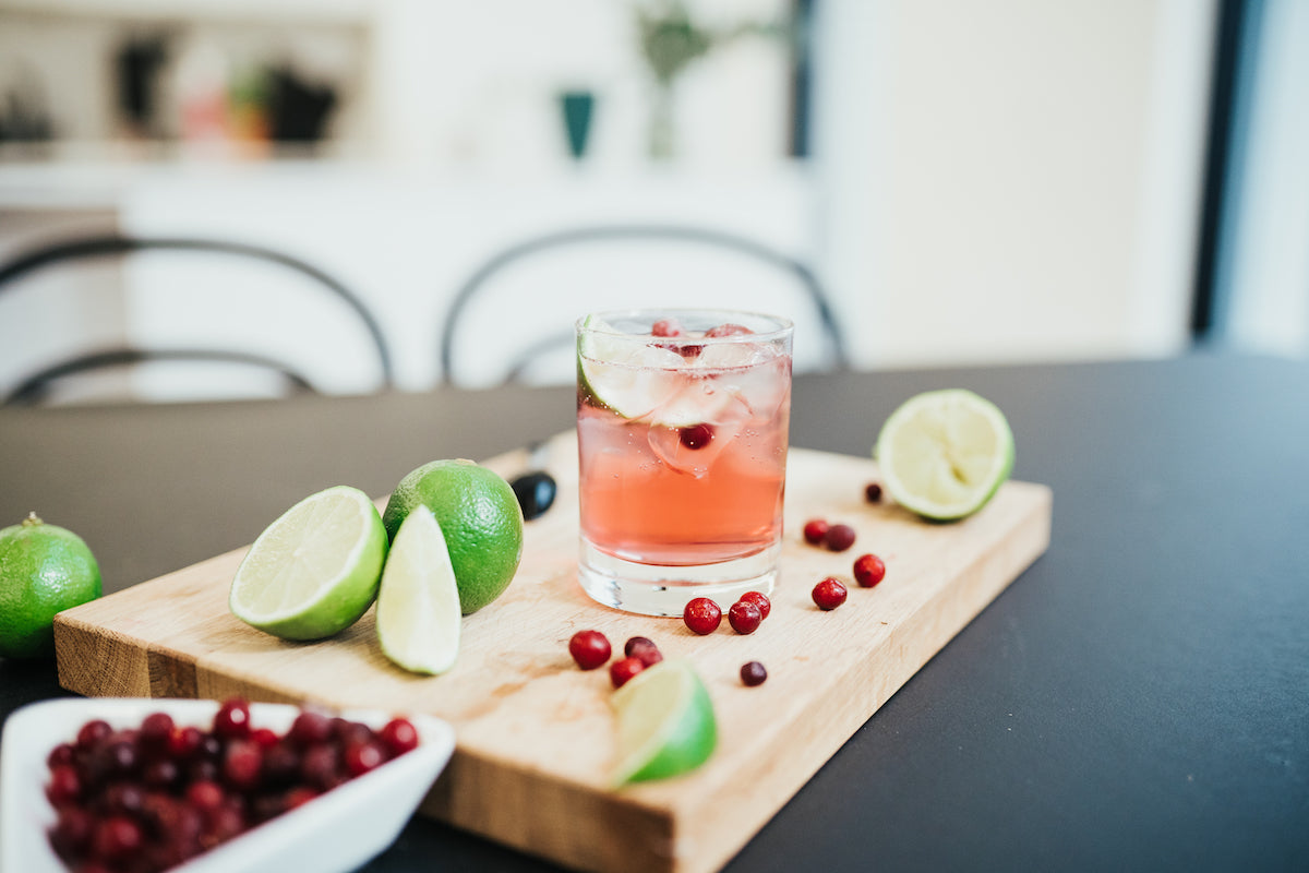 A glass of refreshing Mysoda cranberry lime mocktail on a wooden board with lime wedges and cranberries spread around