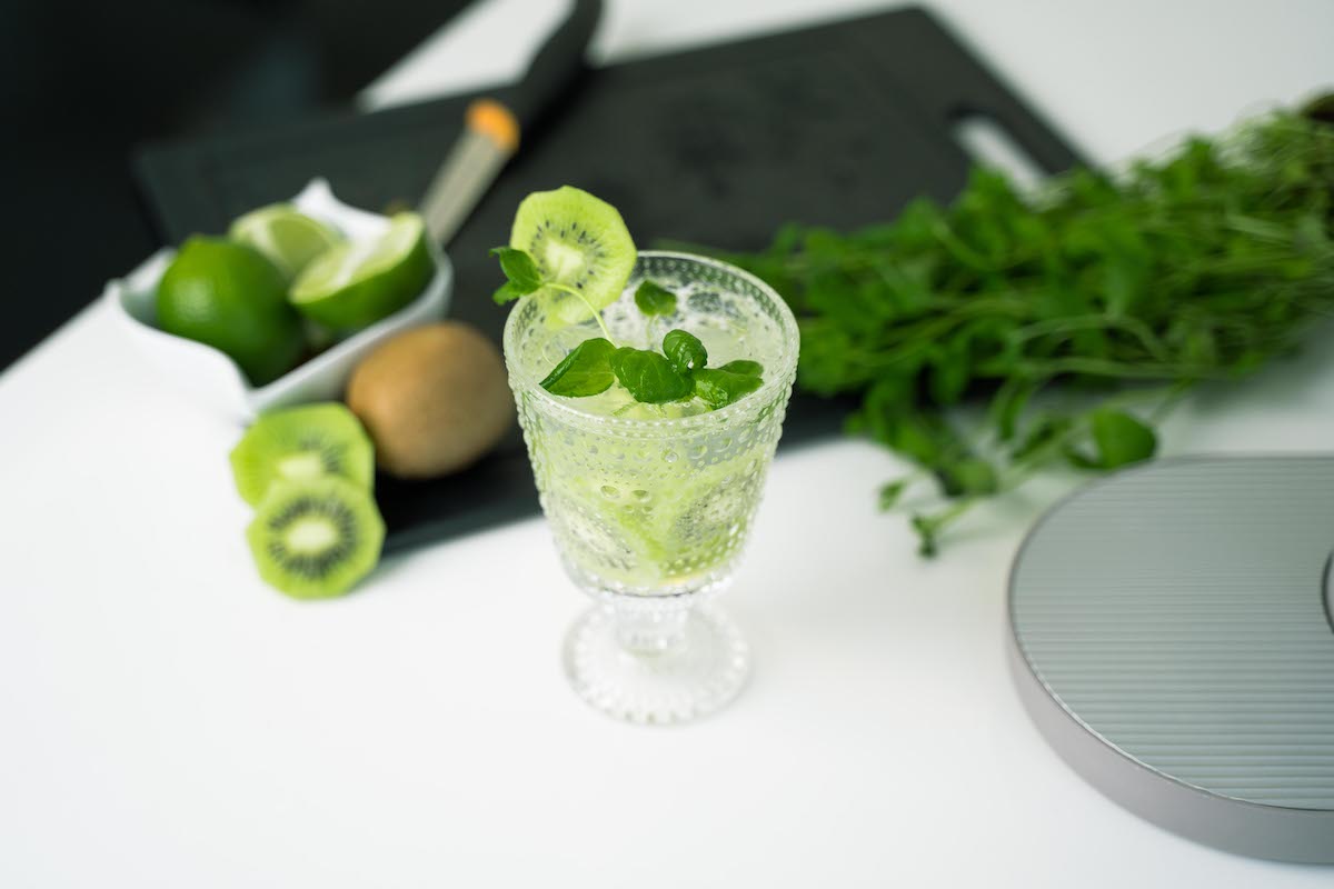 A glass of kiwi mocktail with a cutting board and fruit spread out in the background
