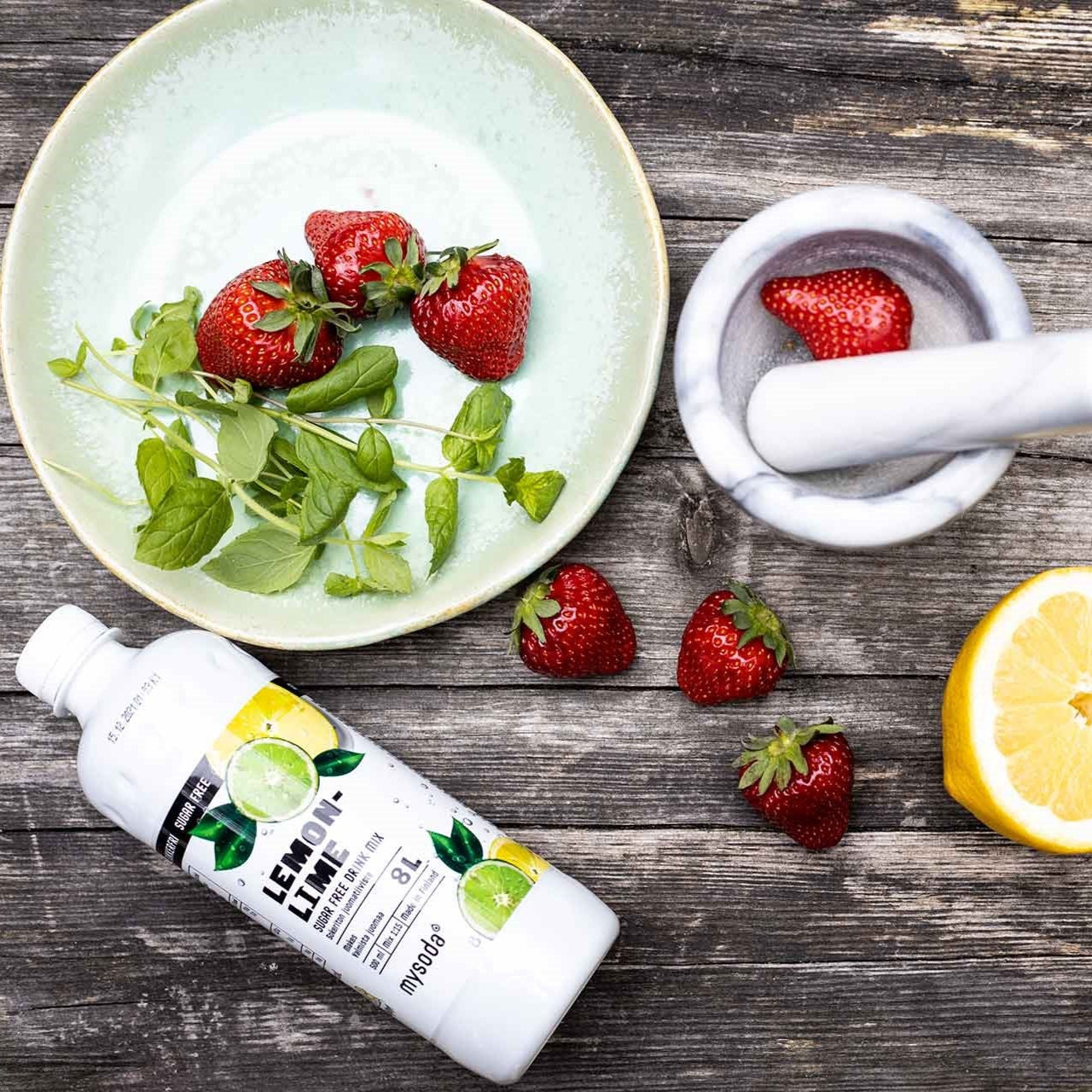 A mix of fruit and herbs as well as a bottle of sugar-free Mysoda drink mix concentrate spread out on a wooden table