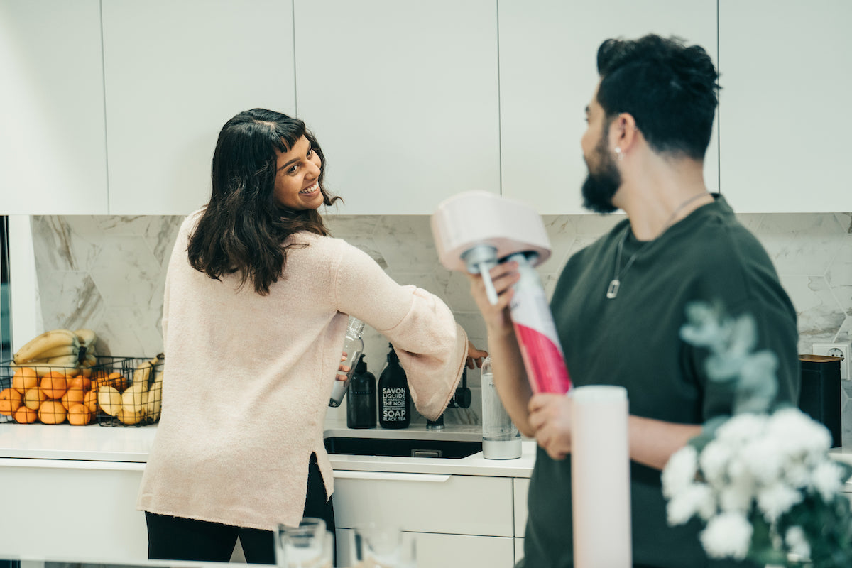 A couple interacting in a white kitchen while the man is changing the co2 cylinder of a pink Mysoda Ruby sparkling water maker