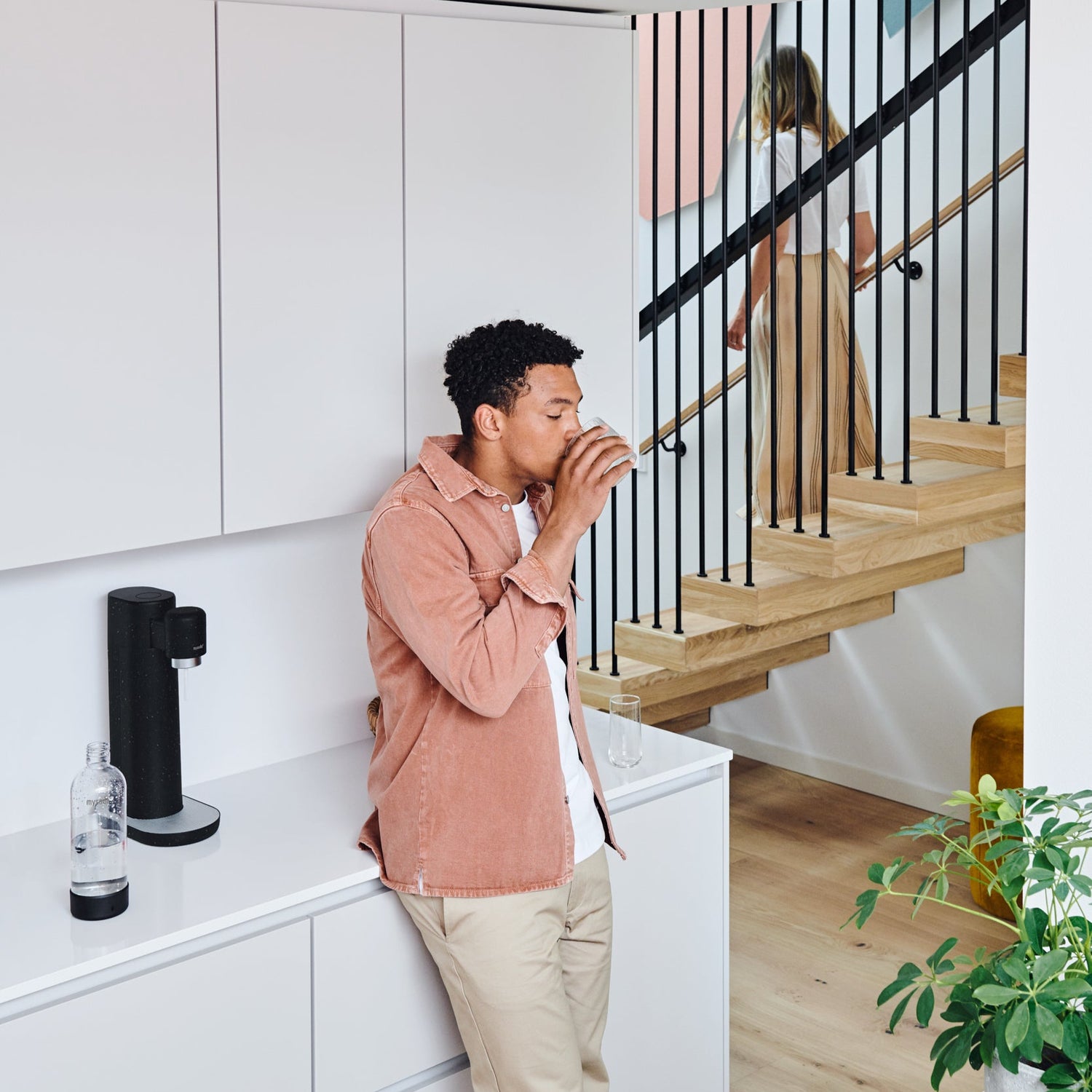 A man drinking sparkling water in a white kitchen with a black Mysoda Toby sparkling water maker standing on the kitchen counter
