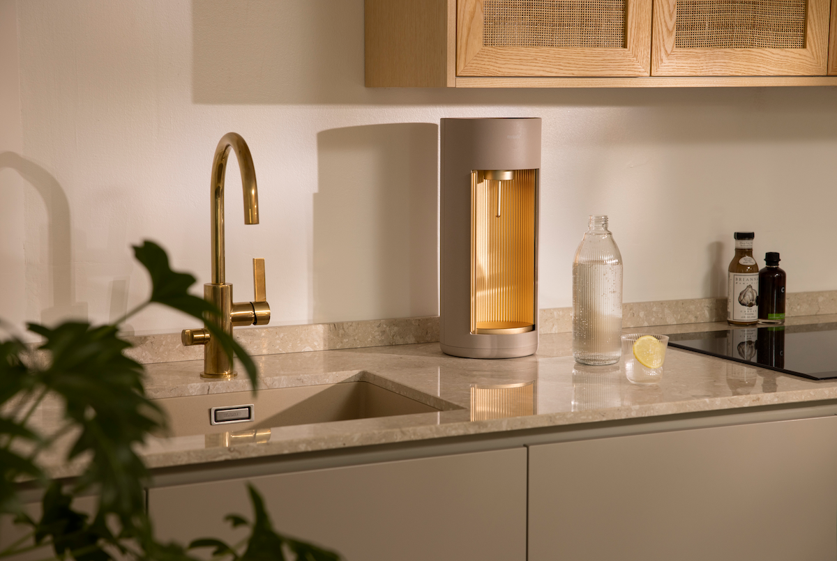 A Glassy sparkling water maker, colour option Latte, a glass bottle and a glass of sparkling water with lemon on a kitchen counter