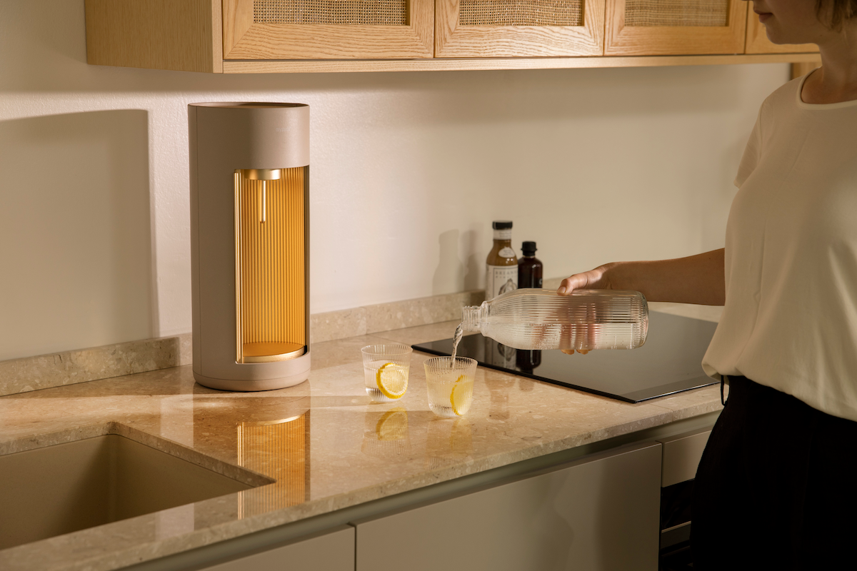 A woman pouring water from a Mysoda glass bottle. A Glassy sparkling water maker, colour option Latte, is visible to the left