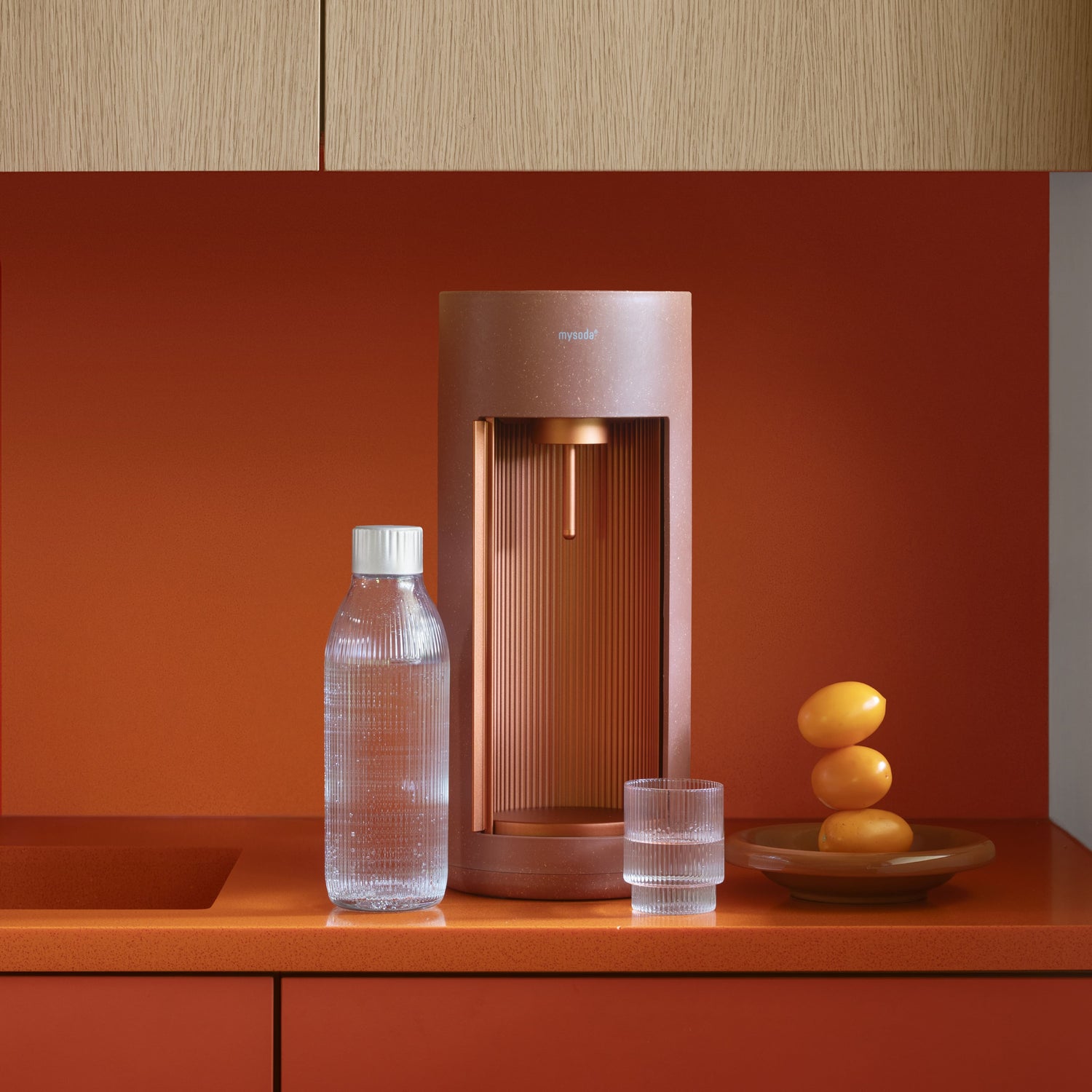 A terracotta-coloured Glassy soda maker with glass bottle and a glass of water on a kitchen counter with orange top and back wall