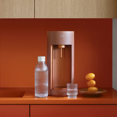 A terracotta-coloured Glassy soda maker with glass bottle and a glass of water on a kitchen counter with orange top and back wall