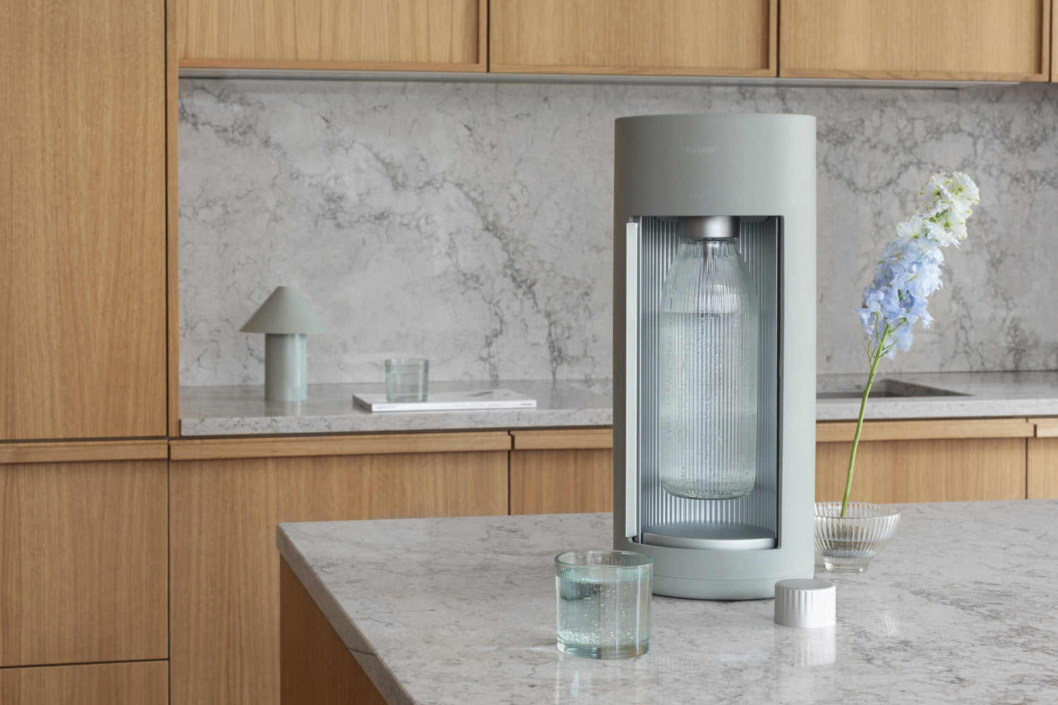 A green Glassy soda maker, colour pigeon, on a kitchen island with lightgray marble countertop and kitchen cabinets of light wood in the background