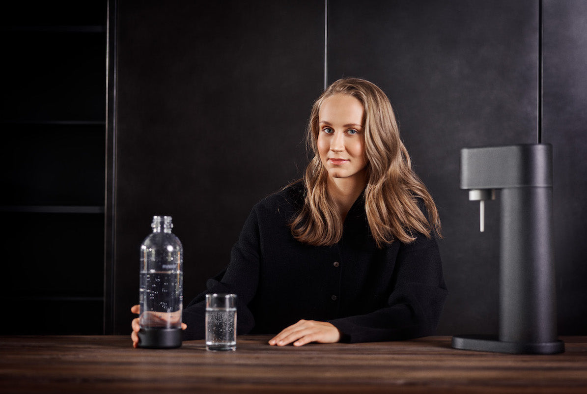 Hurdler Viivi Lehikoinen enjoying fresh sparkling water. A black Woody soda maker is standing on the table to her right and the setting is dominated by black colours