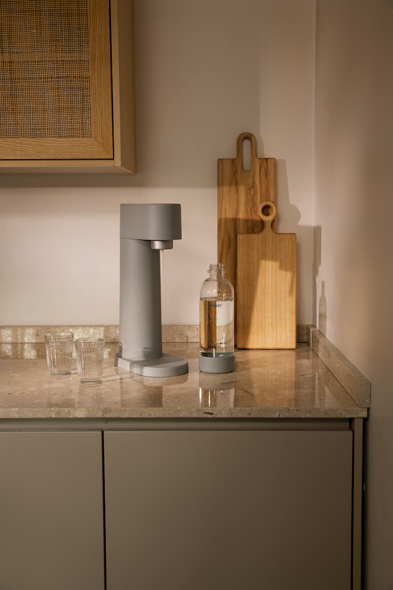A pigeon-coloured Woody soda maker, a water bottle, and two glasses of sparkling water on a kitchen counter