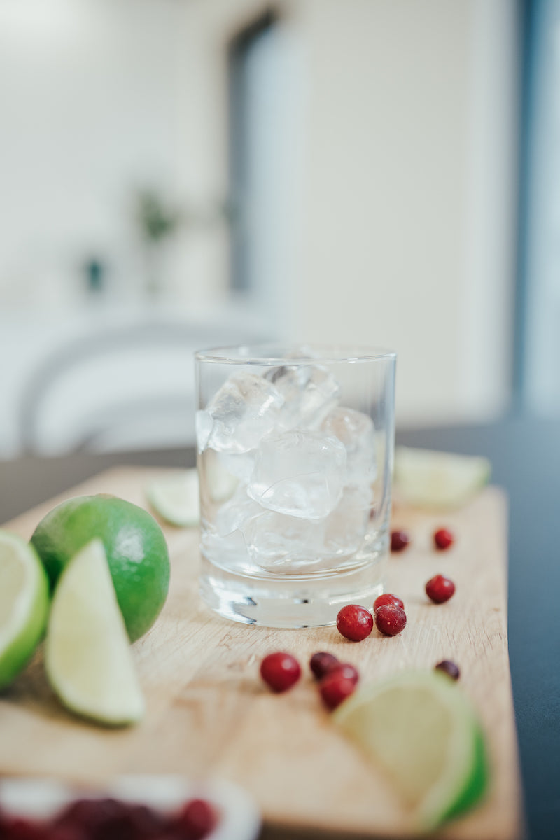 A glass filled with ice cubes surrounded by cranberries and lime wedges on a wooden board