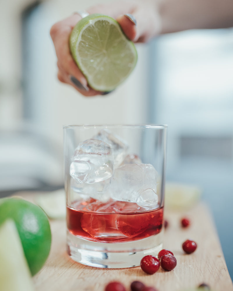 Fresh lime being squeezed into a glass filled with ice cubes and Mysoda cranberry syrup