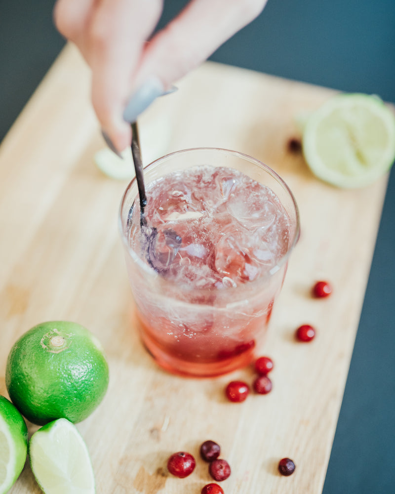 A glass of refreshing Mysoda cranberry lime mocktail on a wooden board with lime wedges and cranberries spread around