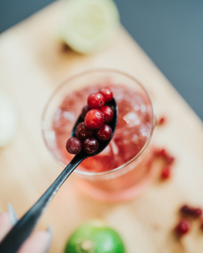 A spoon full of cranberries being added to a glass of bubbly mocktail