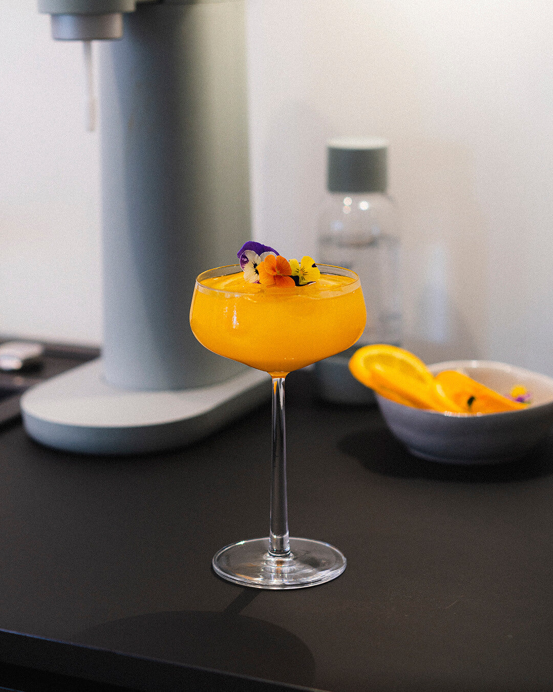 A glass of colourful mango tangerine sparkling mocktail drink with a pigeon Toby sparkling water maker in the background