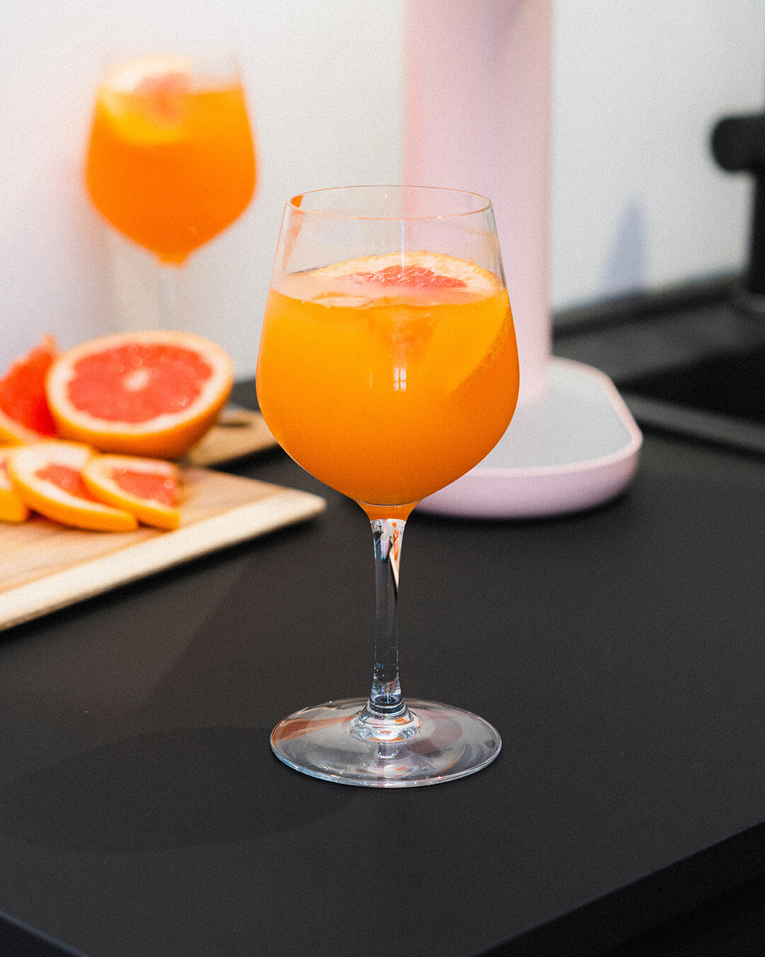 A tall glass of colourful pineapple grapefruit mocktail on a black kitchen counter with Mysoda Ruby sparkling water maker in pink visible in the background