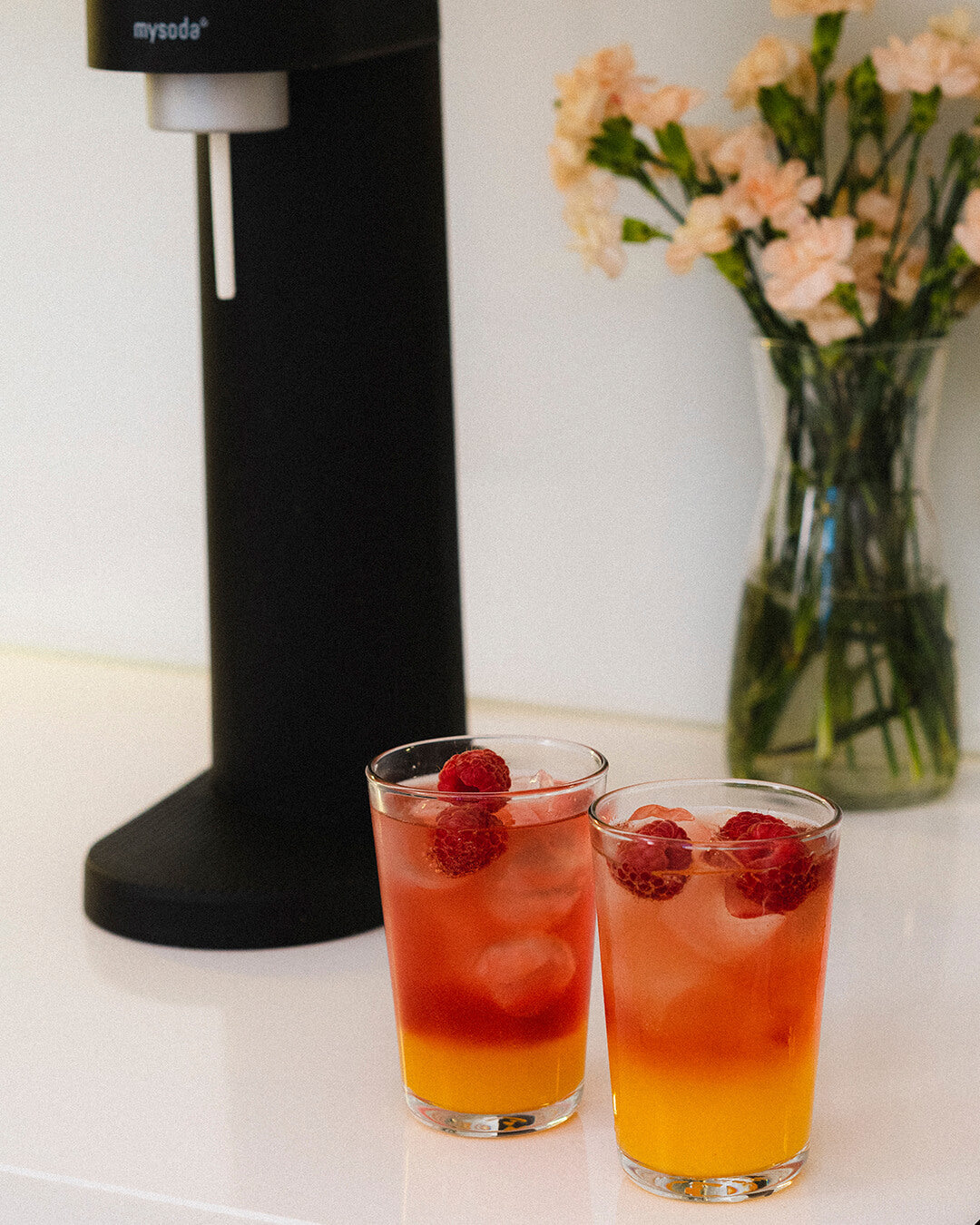 Two glasses of fresh raspberry peach mocktail beverages with a black Woody sparkling water maker visible in the packground