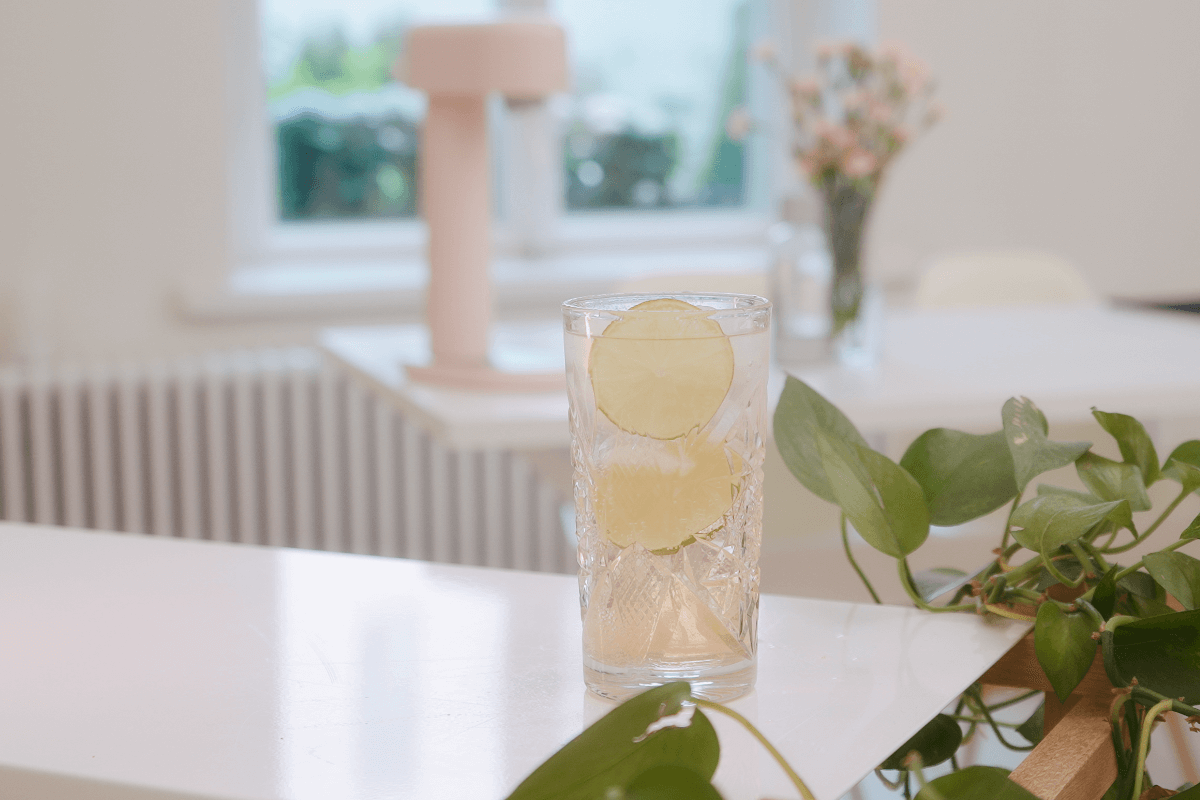 A glass of rhubarb mocktail on a white surface. Across the room one can see a pink Ruby sparkling water maker on a white dinner table