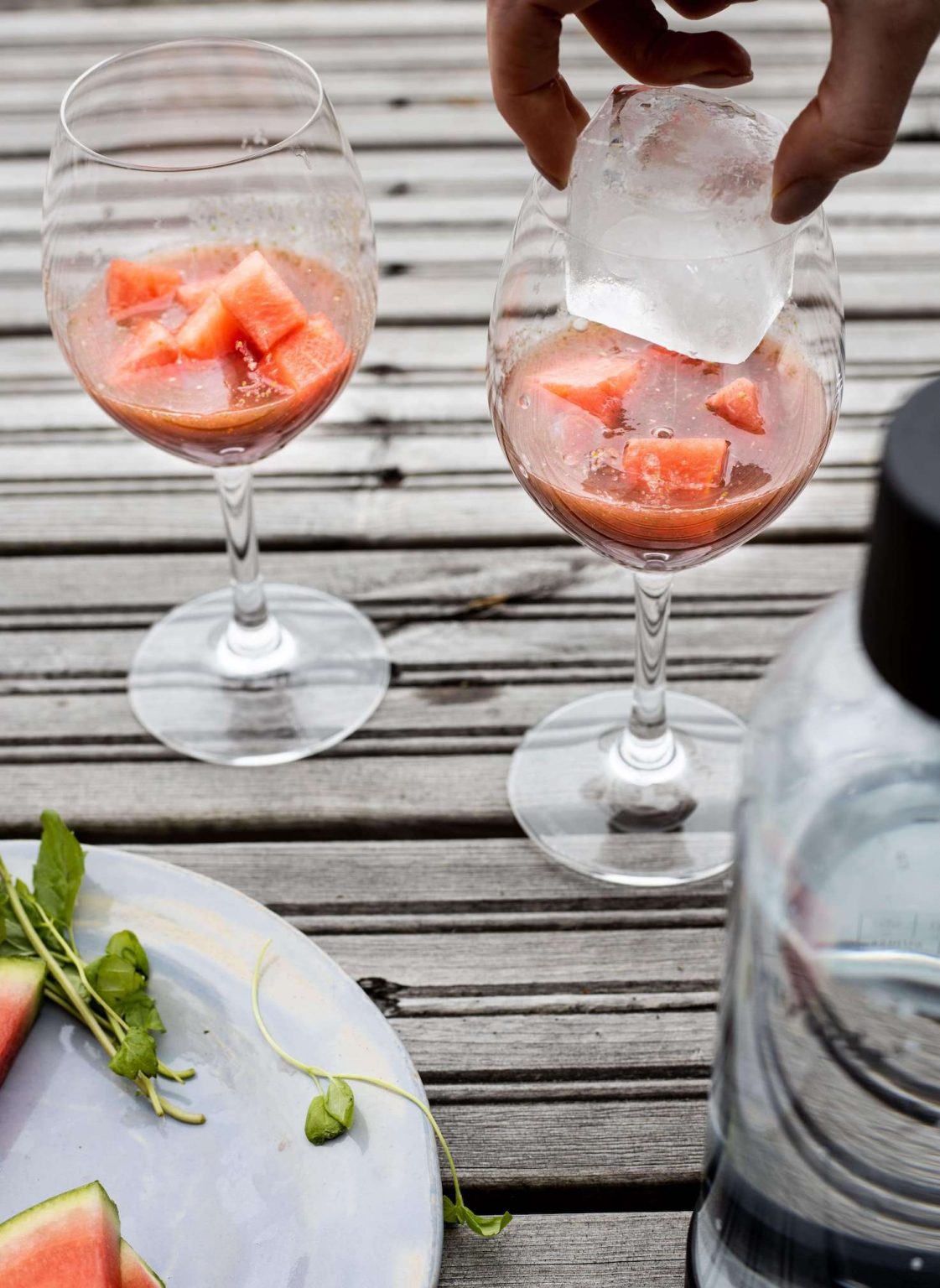 A large ice cube being added to onw of two glasses filled with pieces of watermelon and crushed watermelon