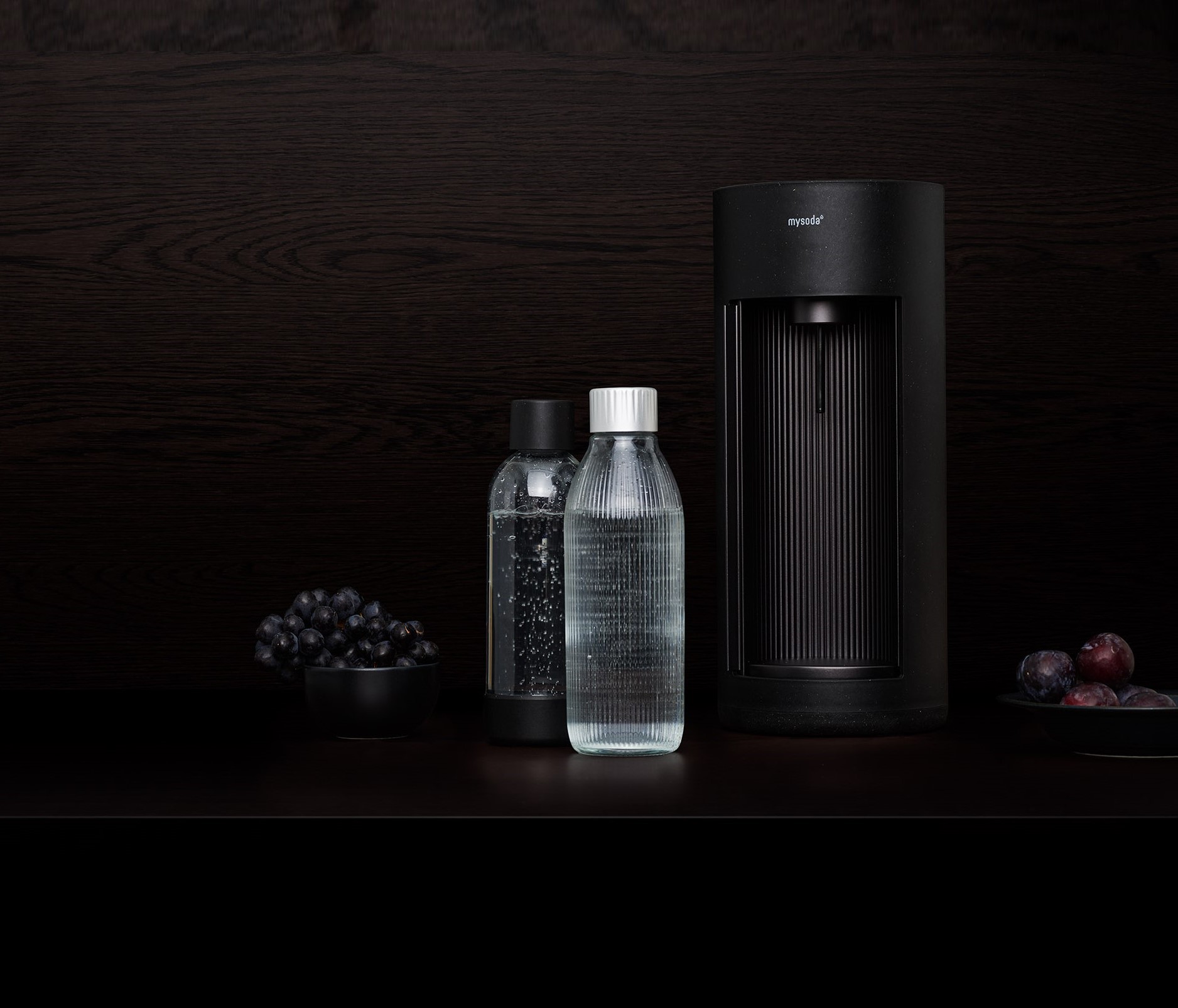 Ein schwarzer Mysoda Glassy Wassersprudler, eine Plastikflasche und eine Glasflasche vor schwarzem Hintergrund