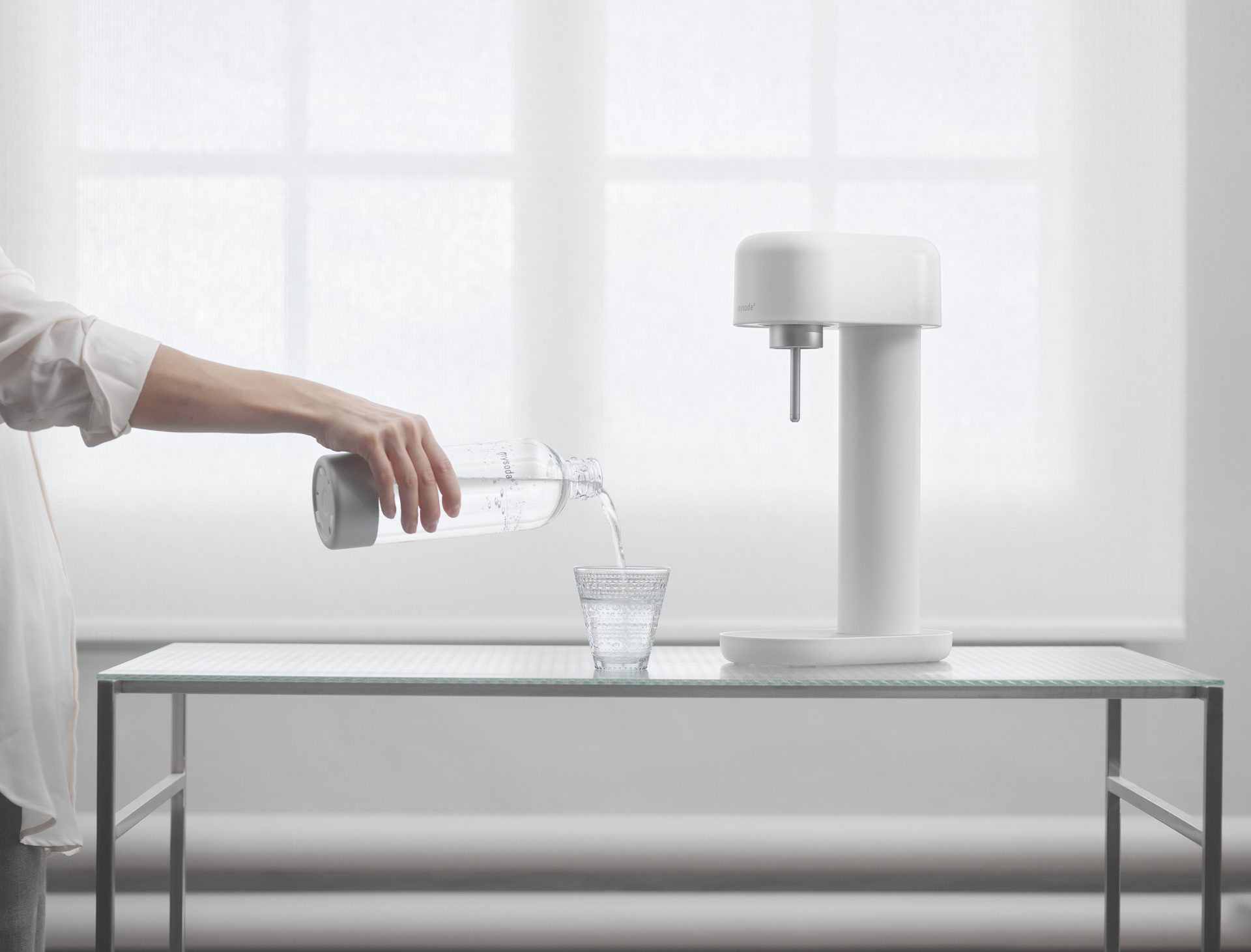 Una macchina per acqua frizzante bianco-argento Ruby 2 davanti a una finestra. Una mano che versa un bicchiere di acqua frizzante.