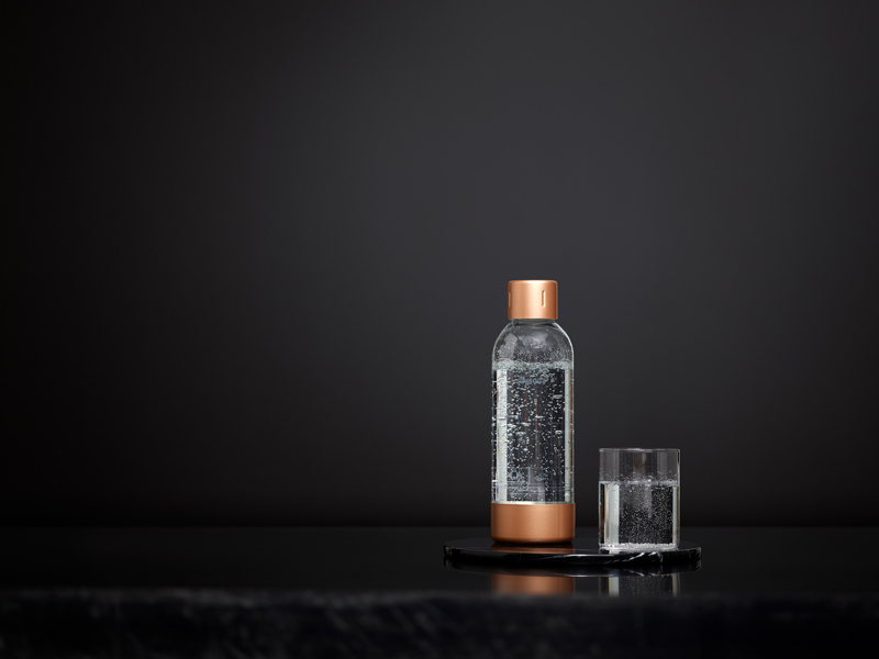A Copper Mysoda premium water bottle and a glass, both filled with bubbly water, in front of a black background