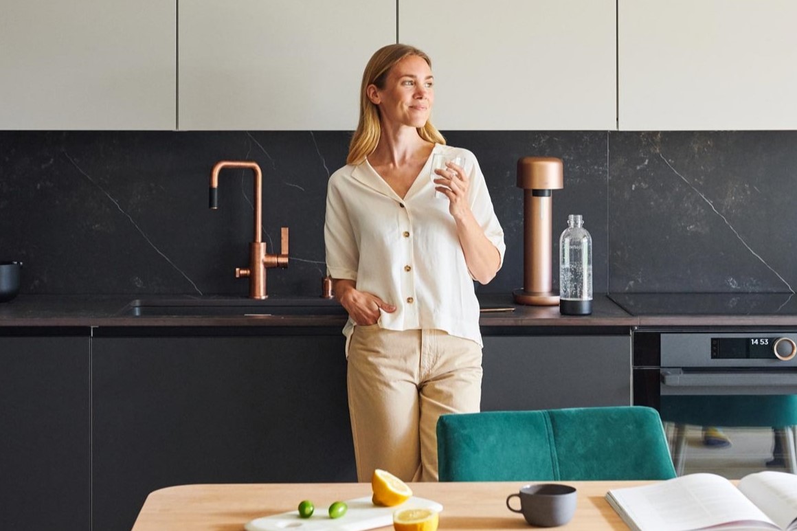 A woman standing next to a Copper Mysoda Ruby sparkling water maker, drinking a glass of sparkling water.