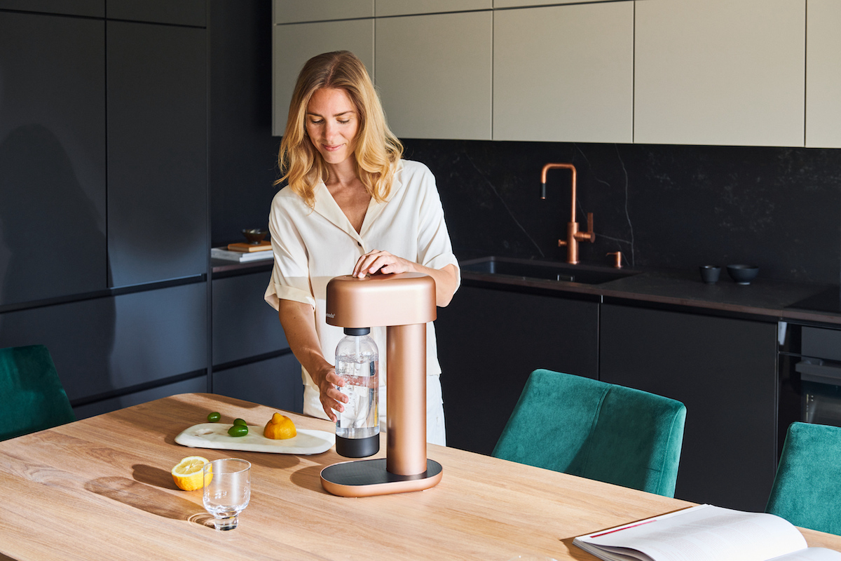 A woman using a copper Mysoda Ruby sparkling water maker in a black kitchen