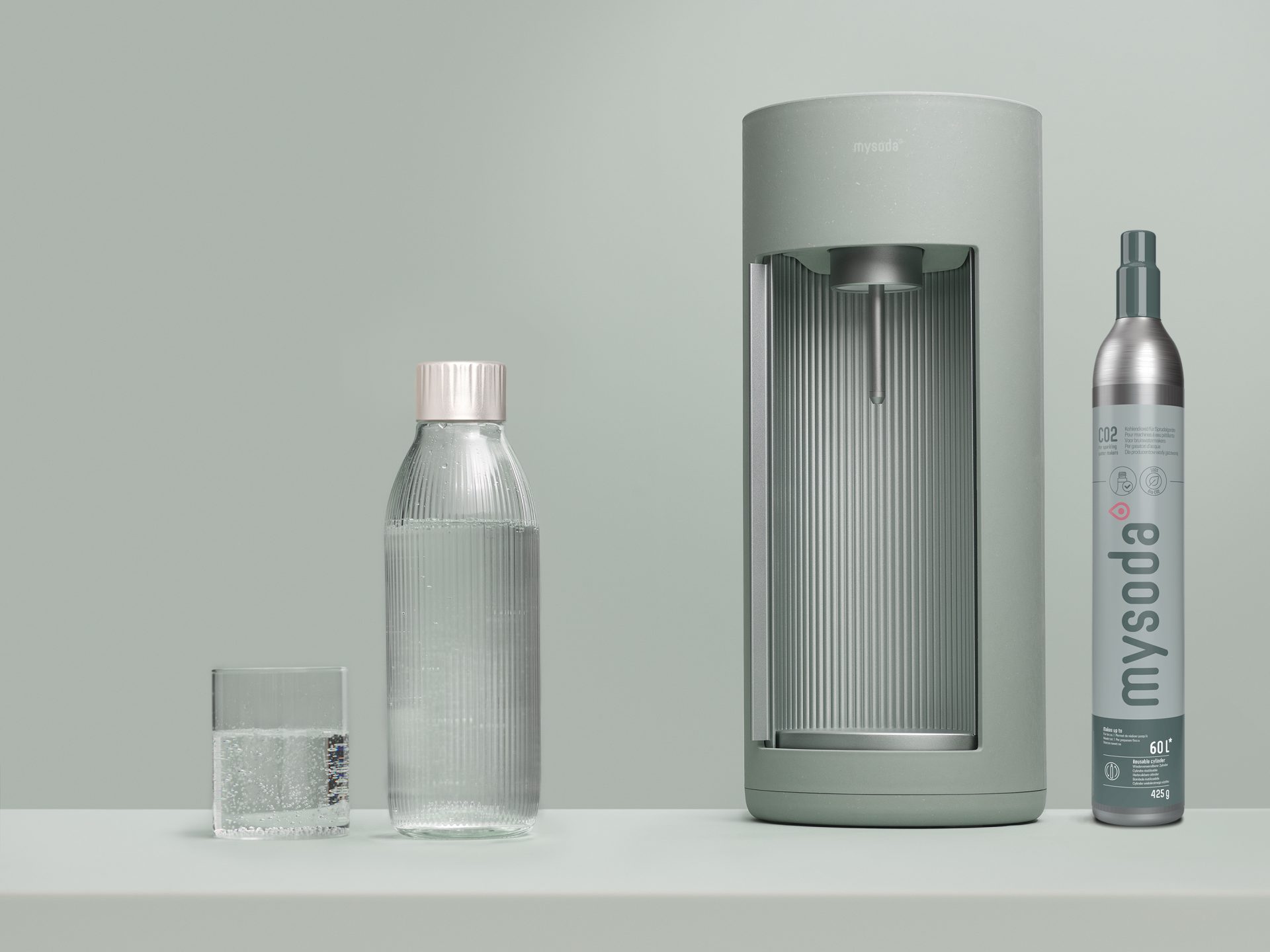 A pigeon coloured Glassy sparkling water maker, a CO2 cylinder with sleeve in similar colours, and a glass bottle in front of tone-to-tone background